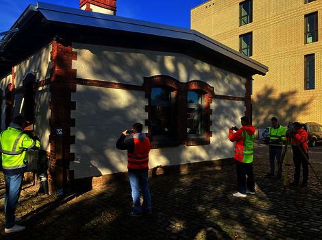 Kleines Haus mit weißer Fassade, dunklem Dach und roten Backstein-Akzenten; mehrere Personen in leuchtend gelben Sicherheitswesten fotografieren das Gebäude im Außenbereich.